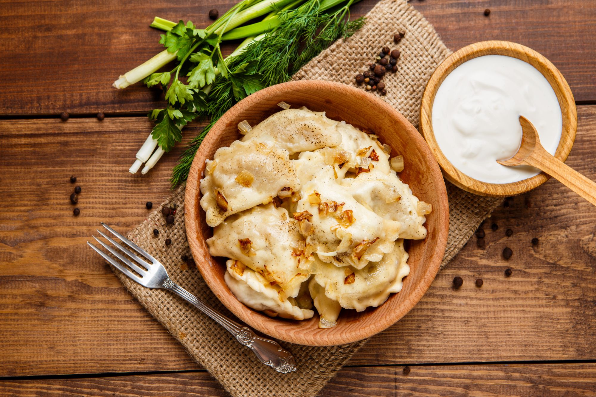 Pierogi w brązowej misce z cebulą, obok śmietana i świeże zioła na drewnianym stole.