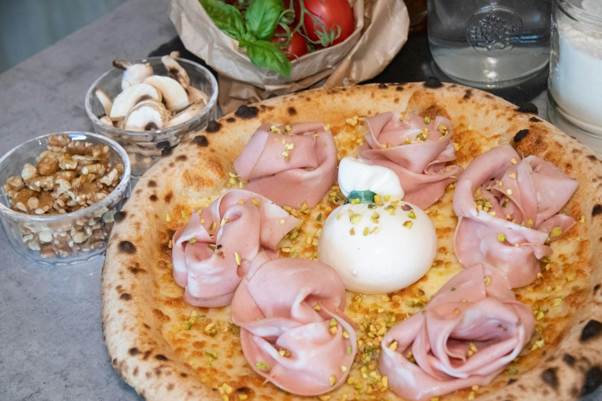 Pizza z serem burrata, szynką, orzechami i grzybami, otoczona ziołami i pomidorami.