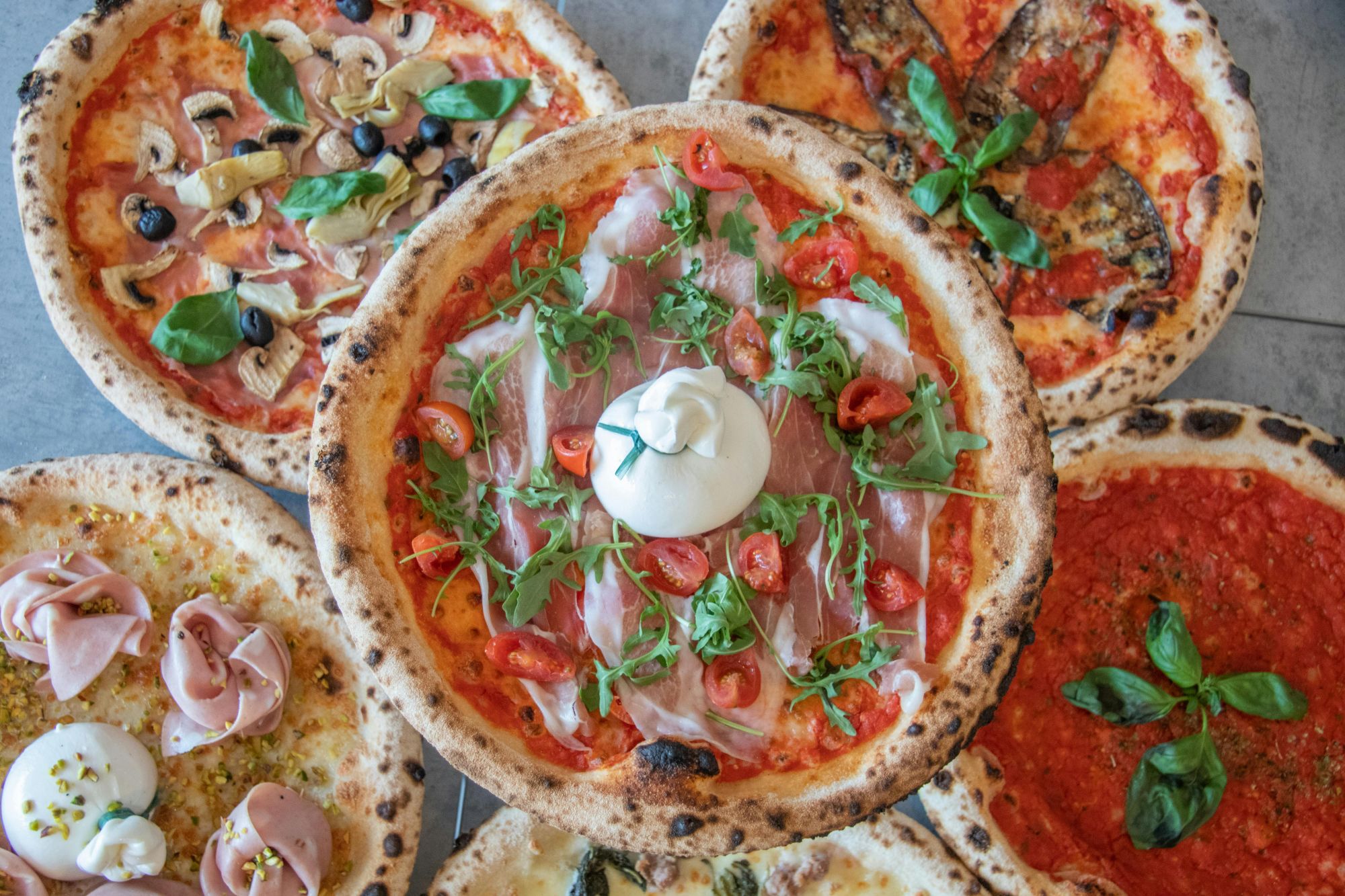 Różnorodne pizze: margherita, z szynką, warzywna, z burratą i dodatkami świeżych ziół.