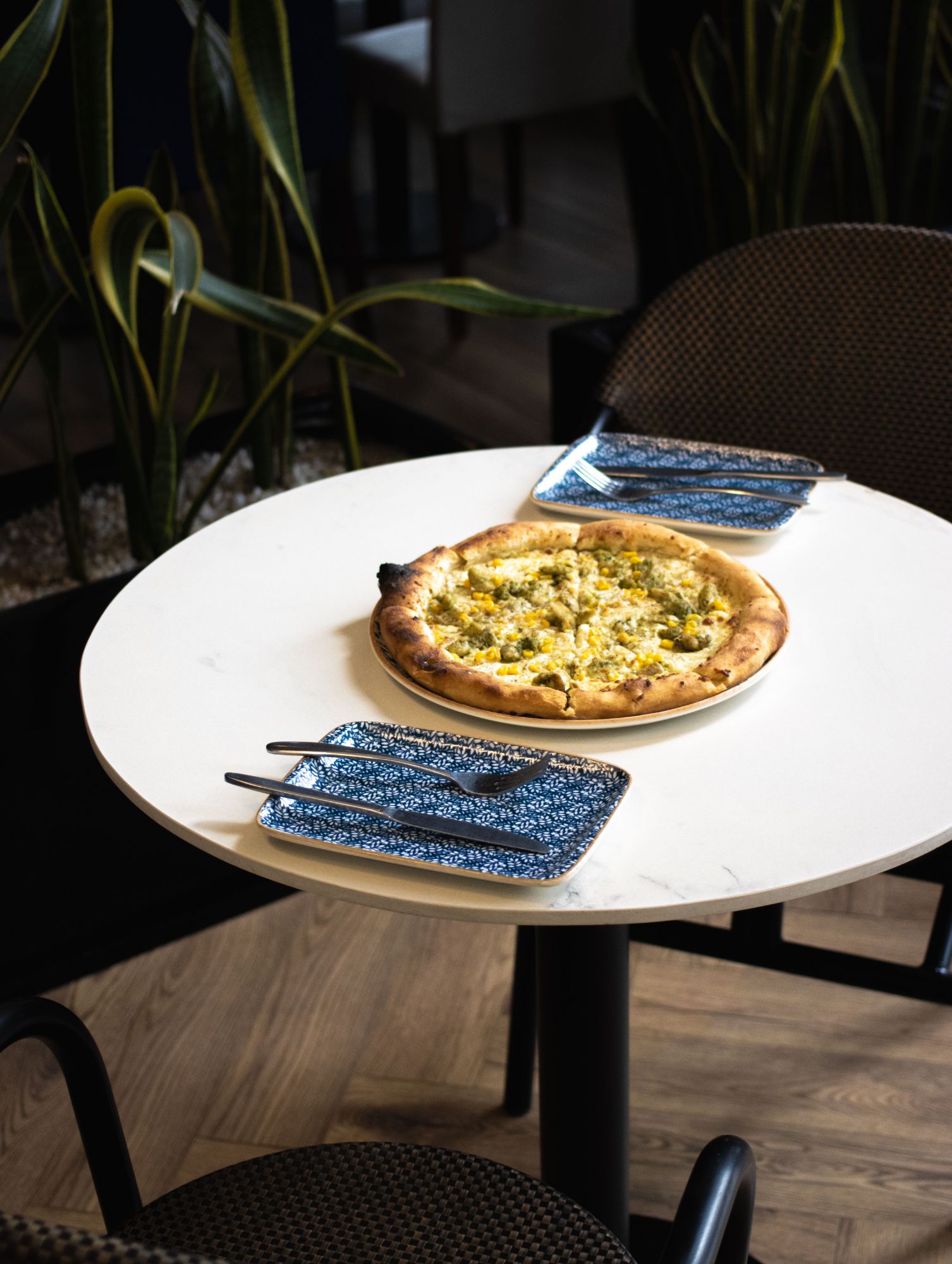 Na stole znajduje się pizza z dodatkami, obok talerze i sztućce, w otoczeniu roślinności.