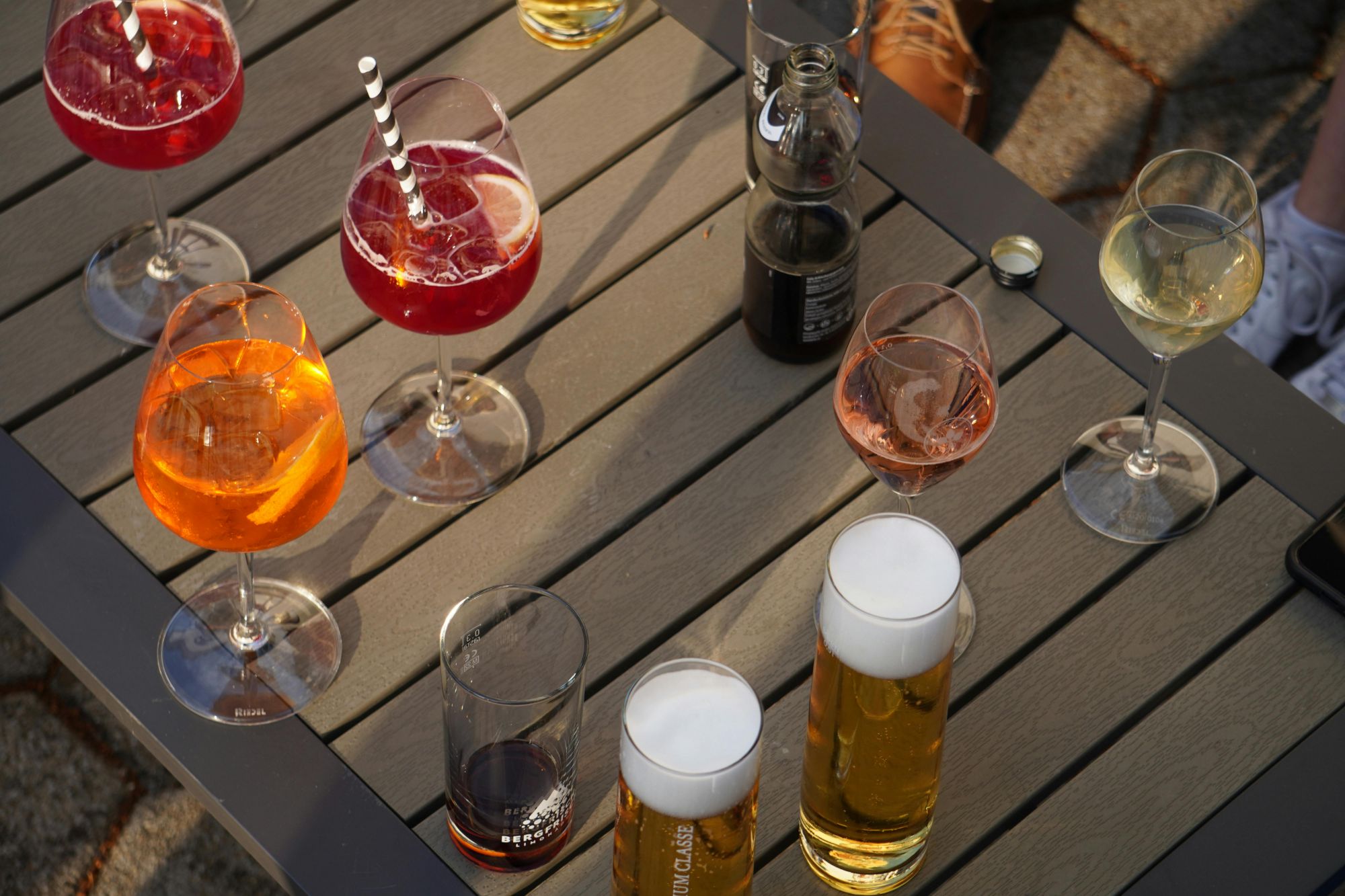 Szeroki wybór drinków i piw na stole, otoczone lampkami i szklankami w słonecznym otoczeniu.