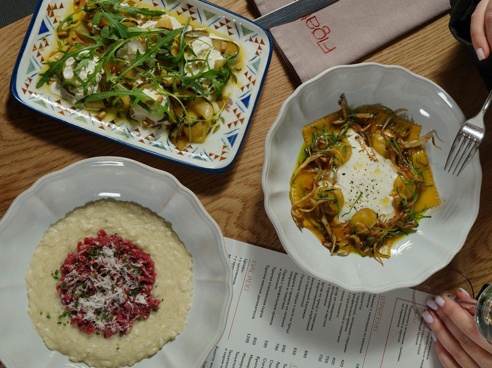 Trzy talerze z potrawami: risotto, sałatka i makaron, na drewnianym stole z menu w tle.
