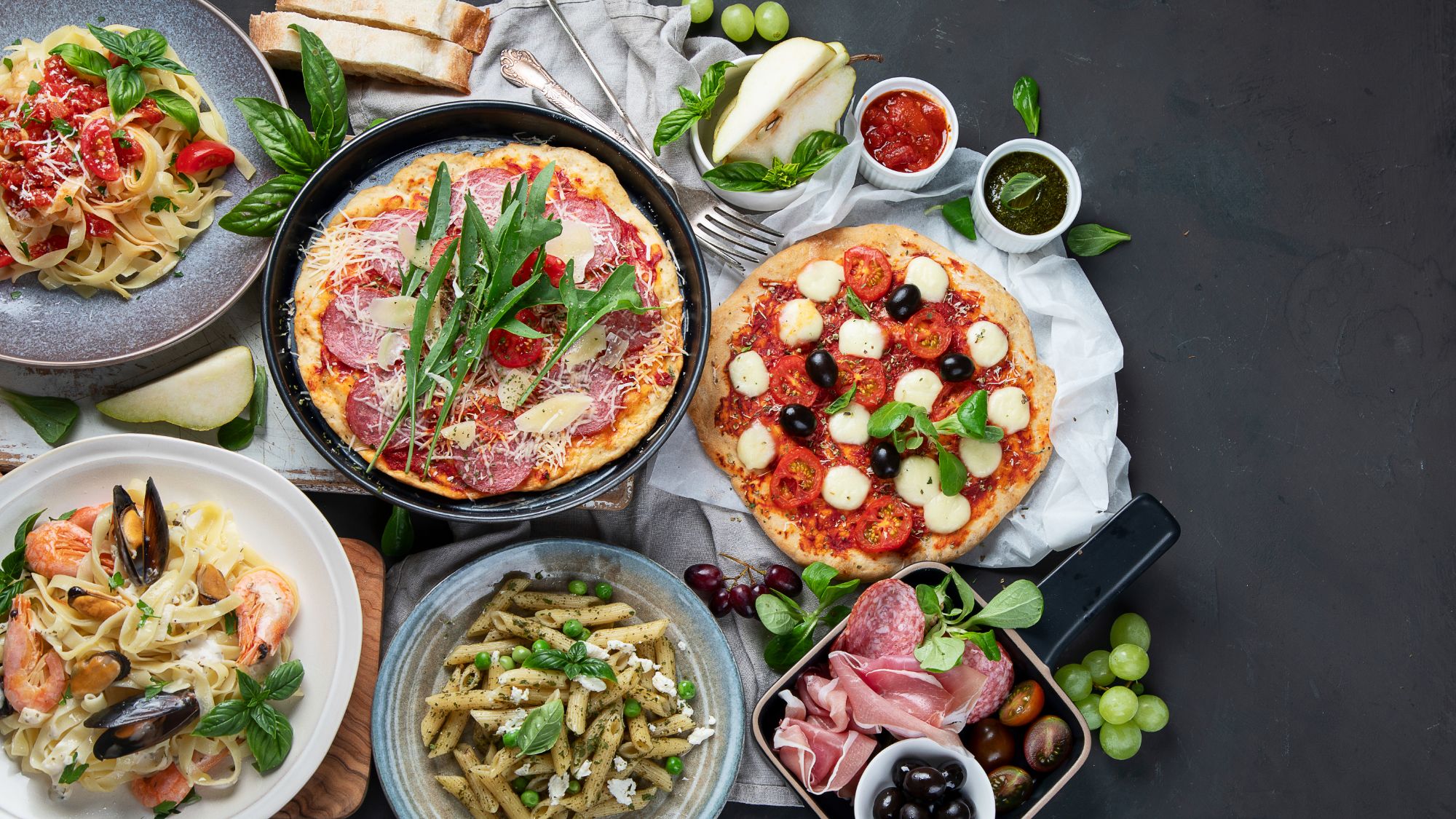 Różnorodne dania włoskie, w tym pizza, makaron i sałatki, podane na stół ze świeżymi ziołami i dodatkami.
