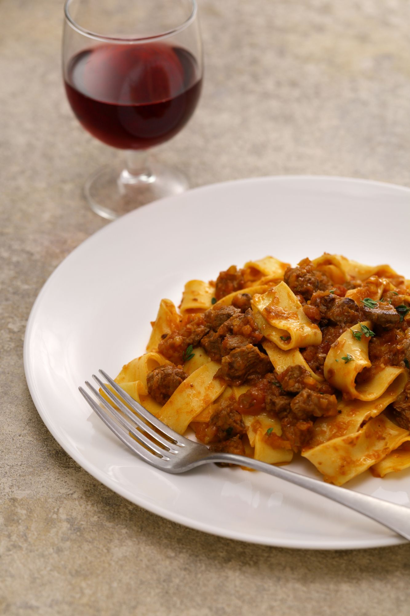 Makaron pappardelle z sosem mięsnym na talerzu, obok kieliszek czerwonego wina.