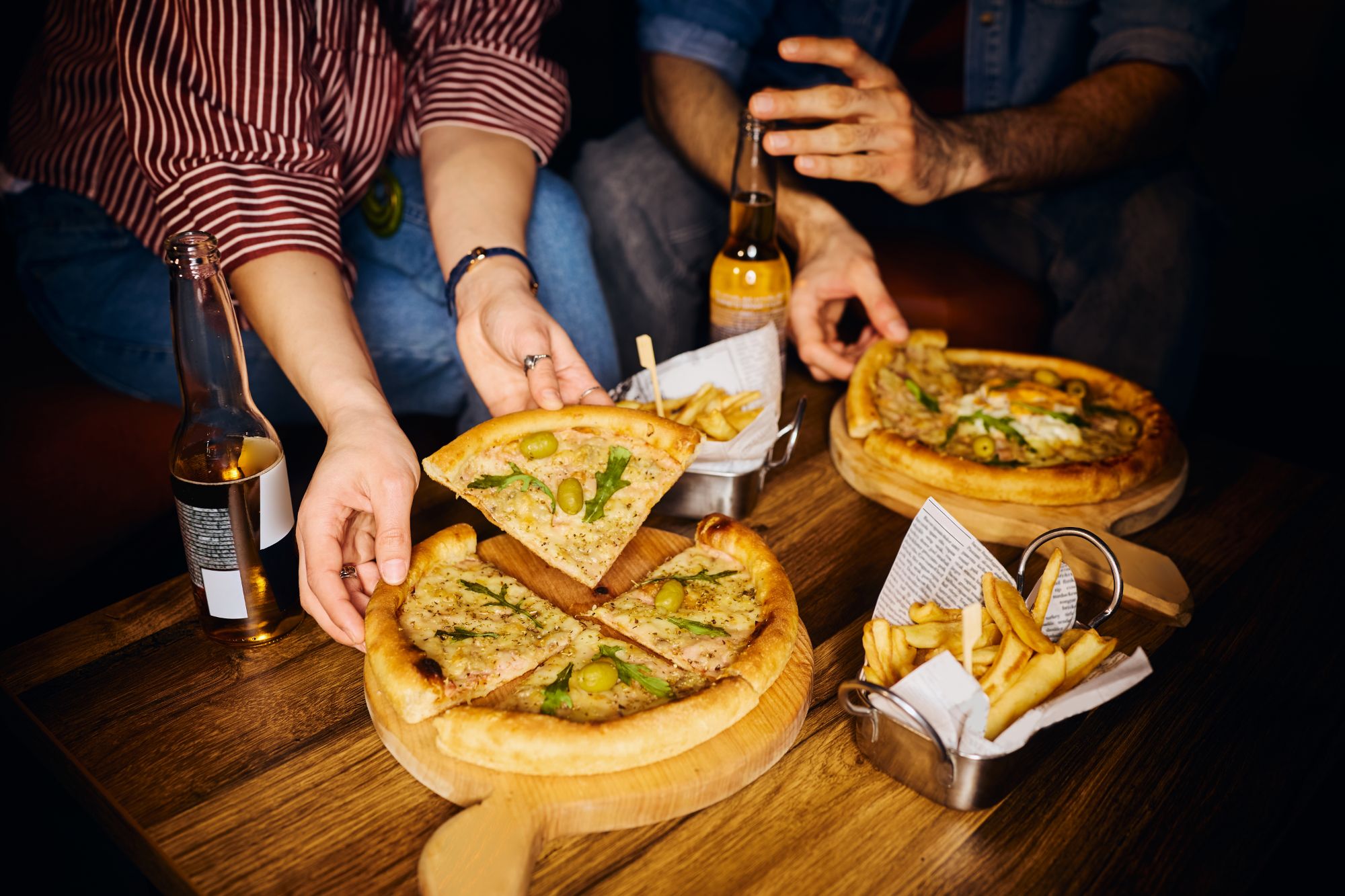 Osoby dzielące się pizzą przy stole, obok frytki i napój w butelce.