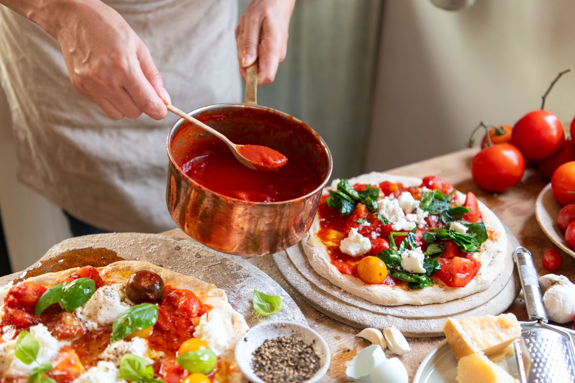 Osoba nalewająca sos pomidorowy na świeżo przygotowaną pizzę z ziołami i warzywami na drewnianym blacie.