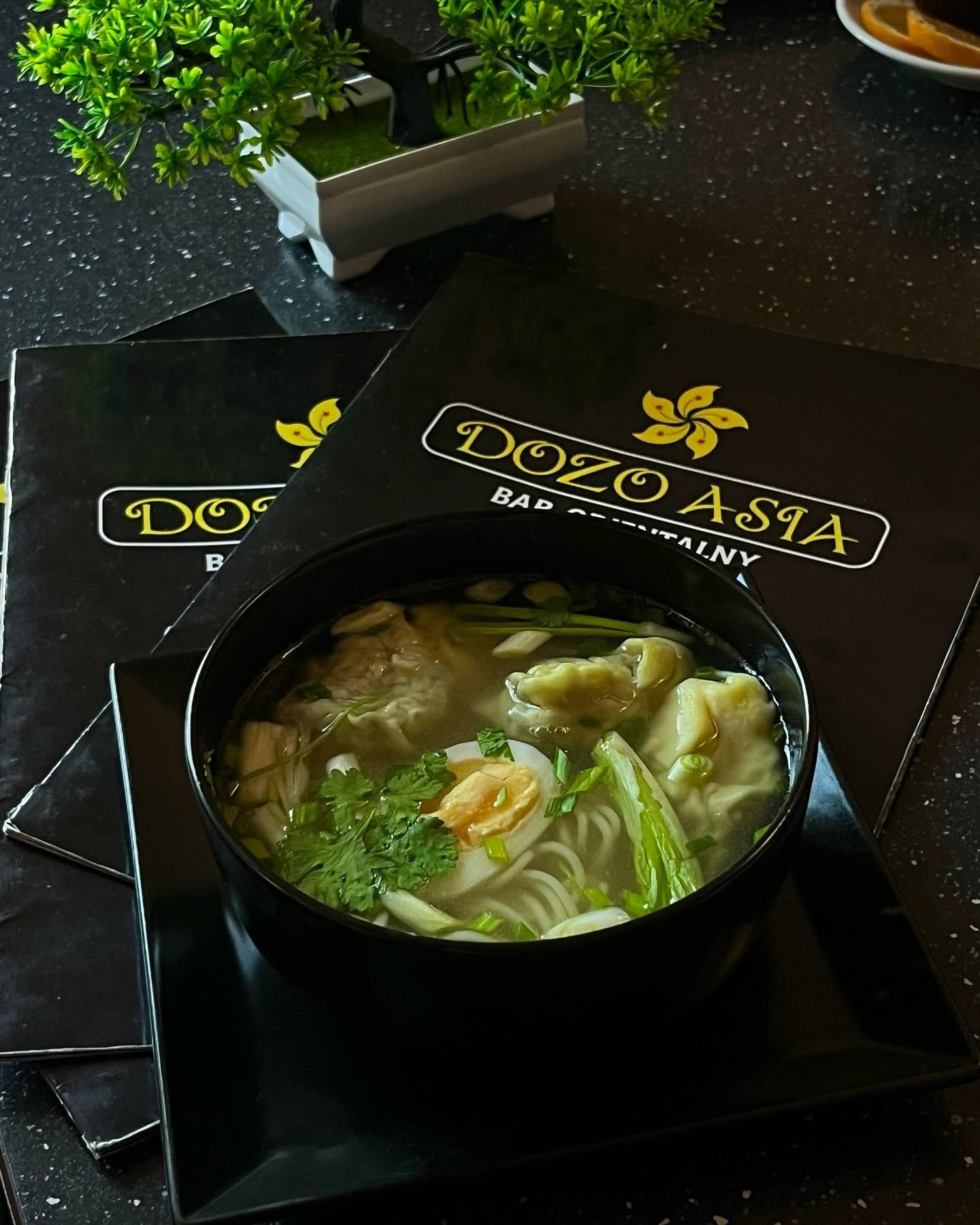 Zupa z makaronem, krewetkami i warzywami w czarnej misce, z menu restauracji Dozo Asia w tle.
