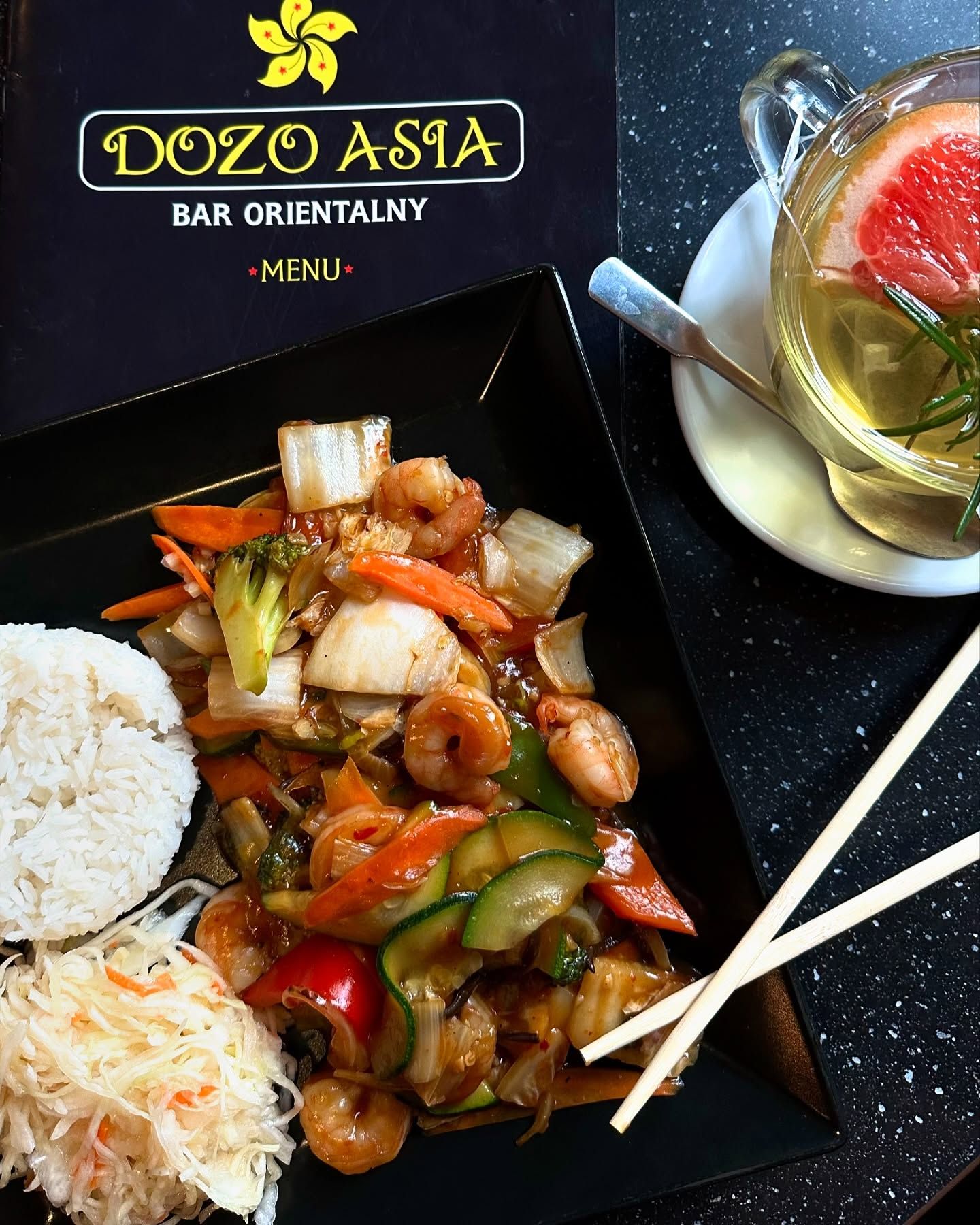 Kolorowy talerz z krewetkami, warzywami, ryżem i herbatą na tle menu restauracji Dozo Asia.