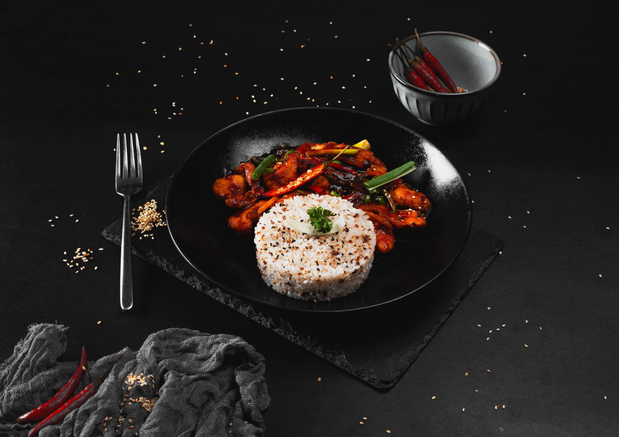 Talerz z daniem: smażone warzywa i kurczak w sosie, podane z ryżem, posypane sezamem, na czarnym tle.