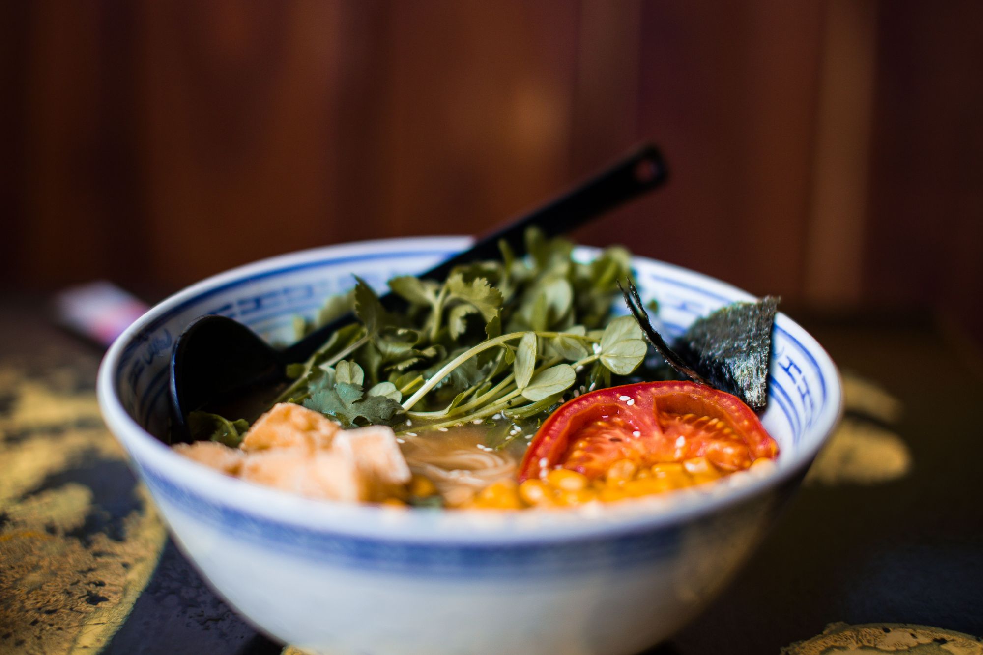 Miska ramen z zieleniną, pomidorami, nori i kawałkami mięsa na ciemnym tle.