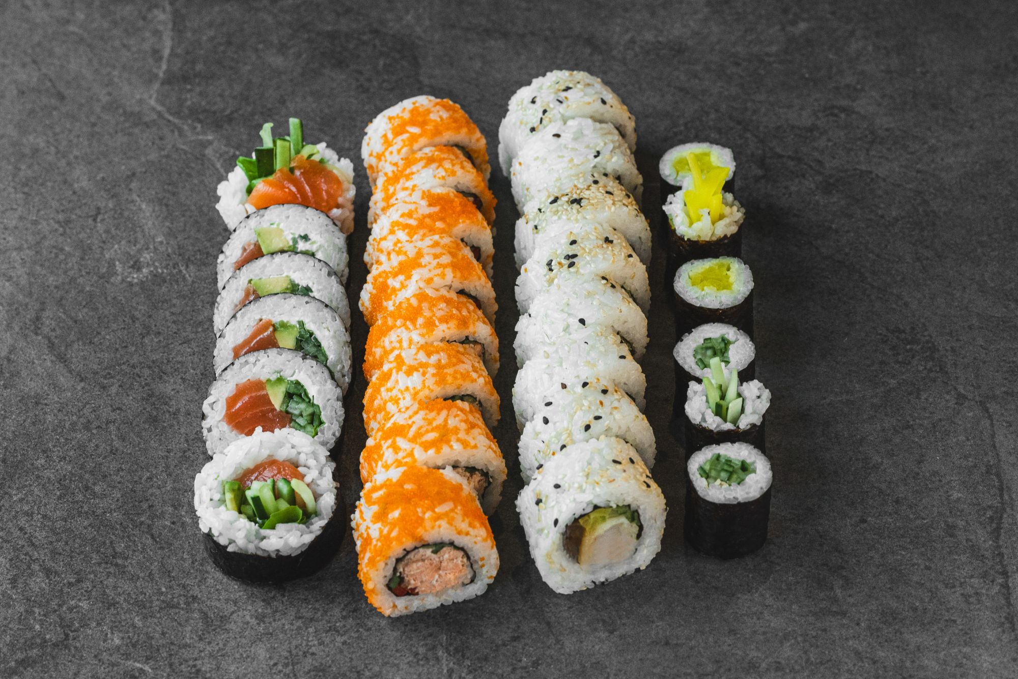 Różne rodzaje sushi na czarnym tle, w tym rolki z łososiem, sałatą i ryżem.