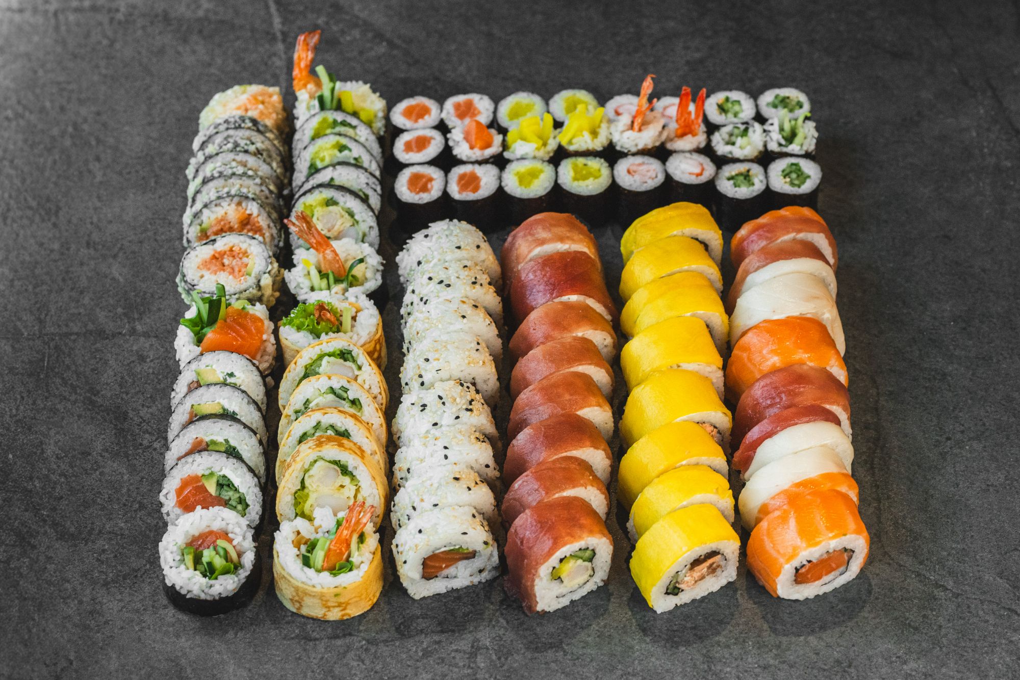 Zestaw różnych rodzajów sushi na ciemnym tle, w tym rolki z łososiem, krewetkami i warzywami.