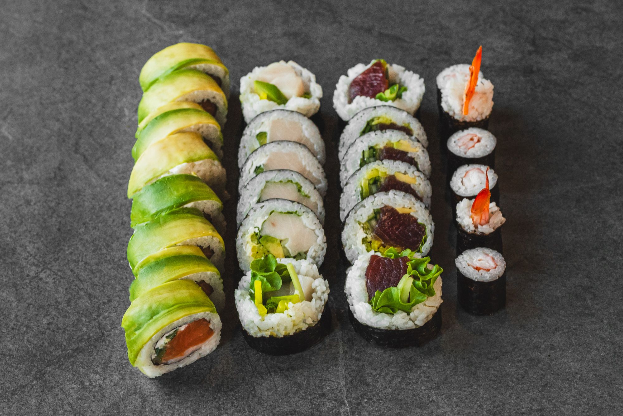 Różnorodne rolki sushi na ciemnym tle, w tym avocado, ryba i warzywa.