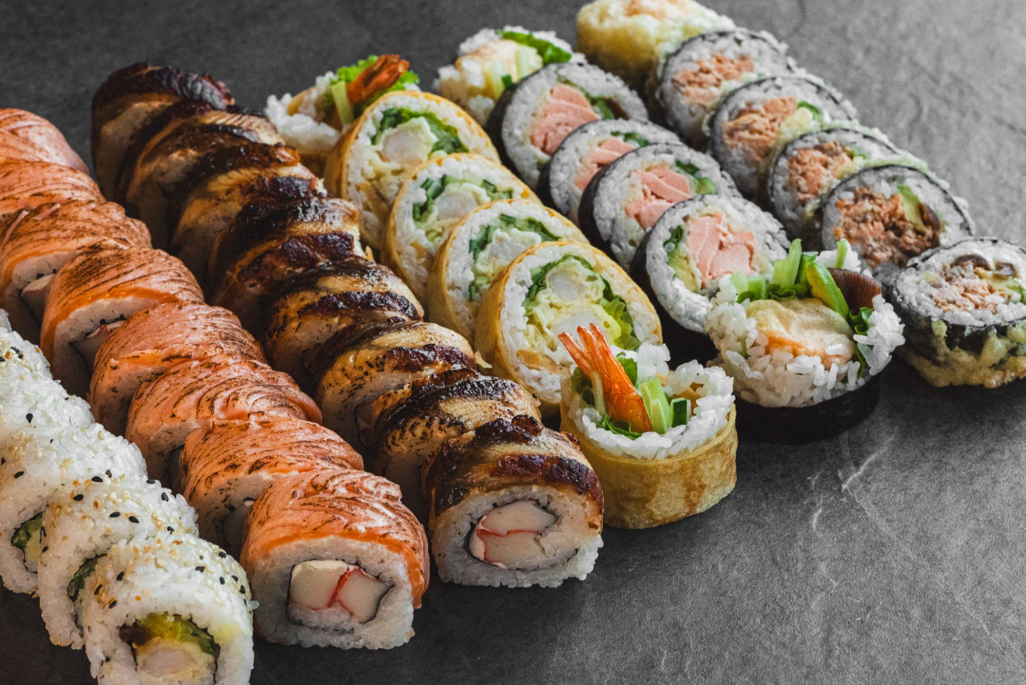 Różnorodne sushi na czarnym tle, w tym rolki z łososiem, krewetkami i warzywami.