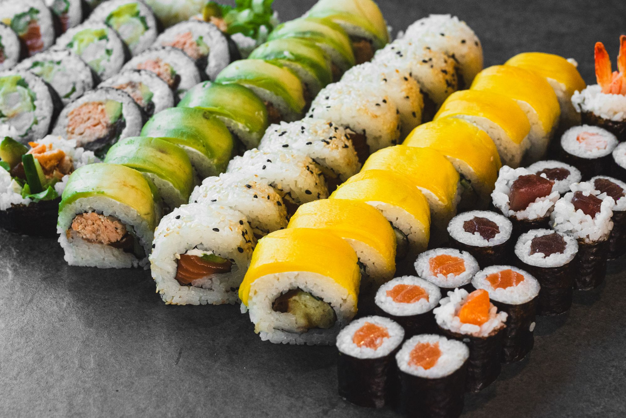 Różne rodzaje sushi, w tym rolki z awokado, mango, tuńczykiem i łososiem, ułożone w estetycznym rzędzie.
