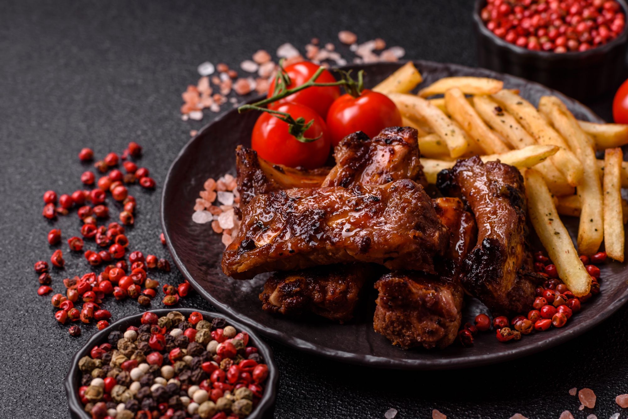 Grillowane żeberka wieprzowe z frytkami i pomidorkami cherry, udekorowane kolorowym pieprzem na czarnym tle.