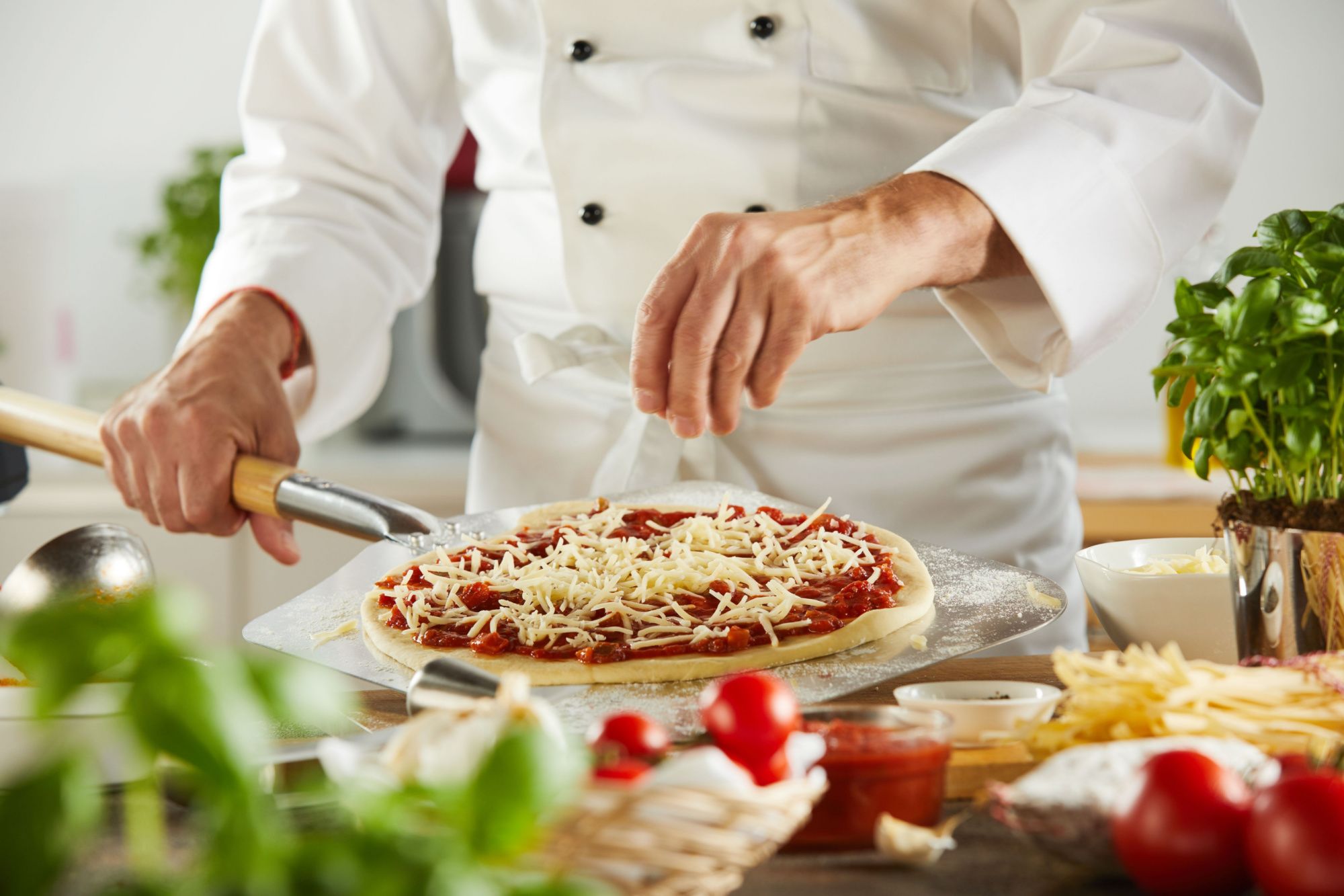 Kucharz posypuje serem startym na pizzy, przygotowując posiłek w kuchni pełnej świeżych składników.