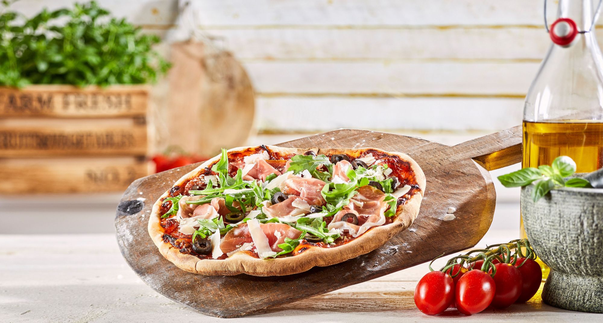 Pizza z szynką i rukolą na desce do krojenia, obok oliwa z oliwek i świeże pomidory.