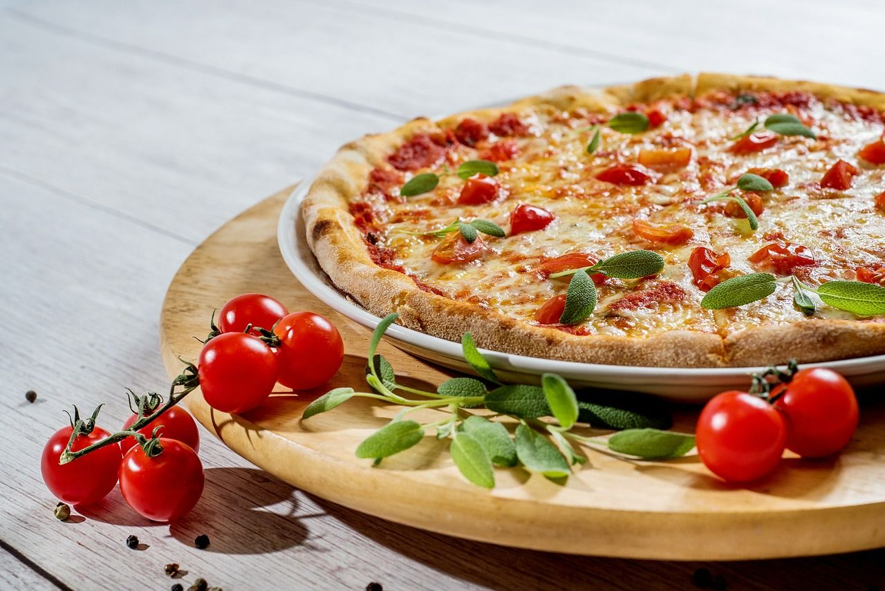 Pyszna pizza z serem i pomidorkami, podana na drewnianym talerzu, obok świeże pomidory i liście szałwii.