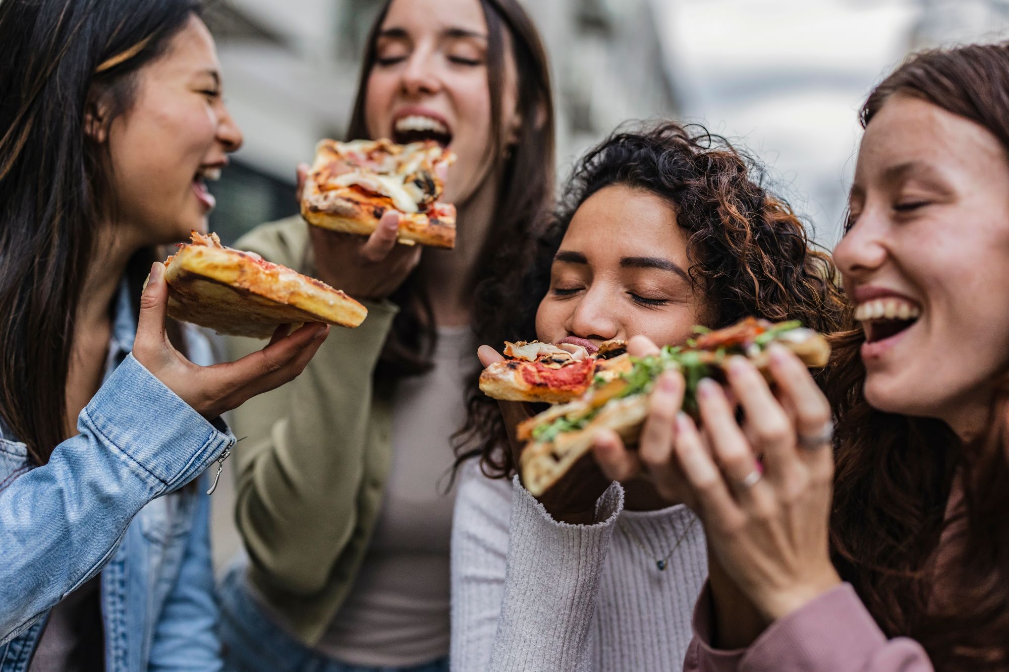Cztery kobiety cieszą się pizzą na zewnątrz, uśmiechając się i dzieląc się jedzeniem.
