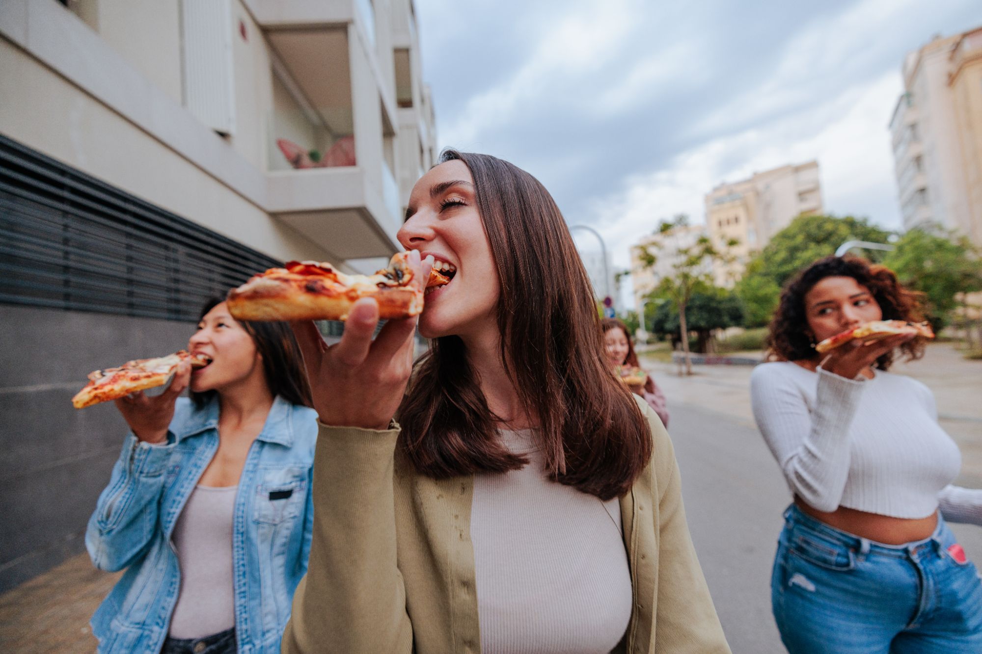 Grupa trzech kobiet spacerujących po mieście, jedzących pizzę.