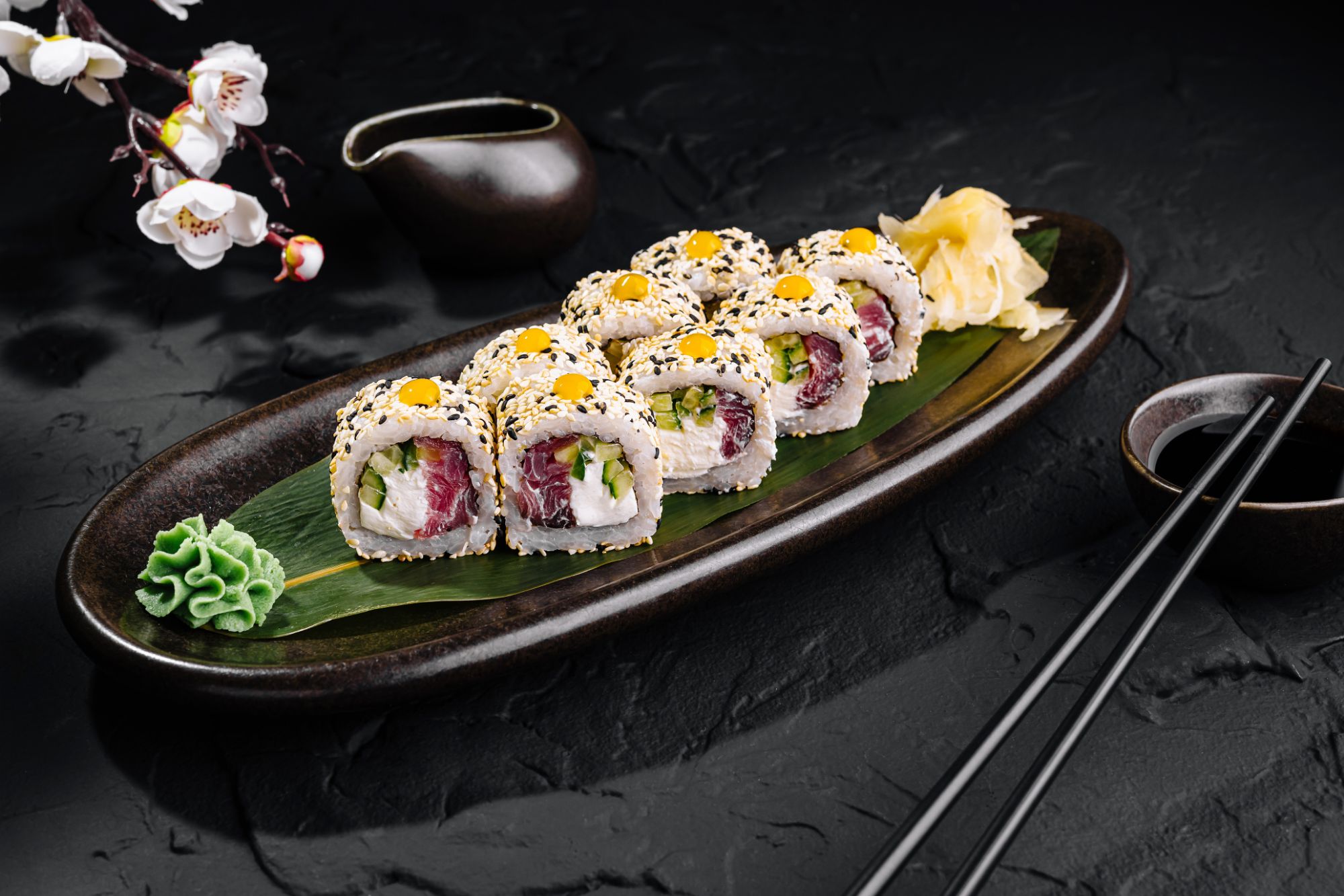 Sushi na talerzu, z ryżem, warzywami, sezamem i dodatkami, otoczone czarnym tłem i kwiatami.