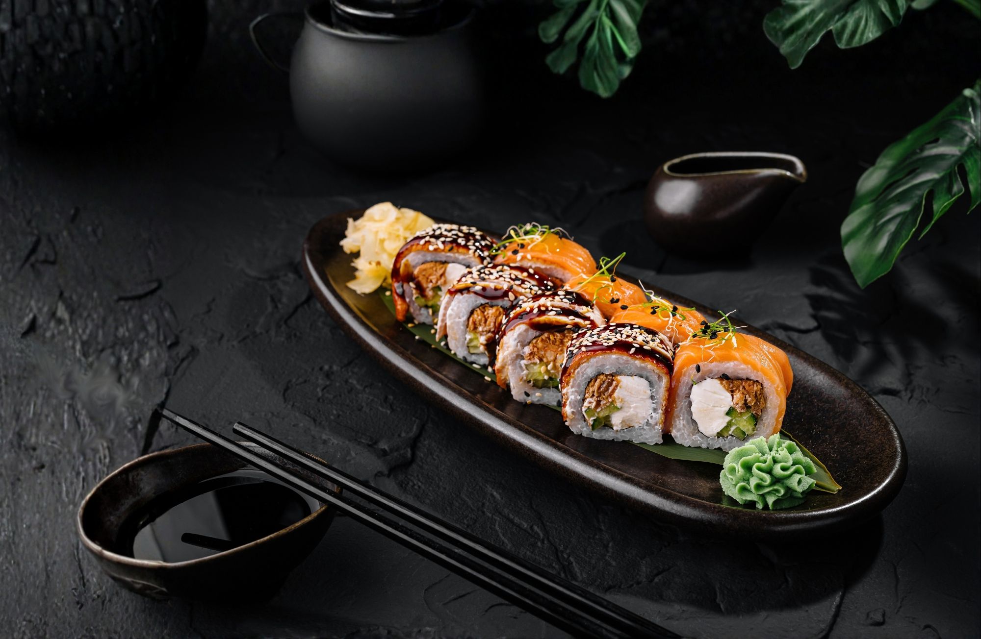Sushi na czarnym talerzu, z rzodkiewką, wasabi oraz sosem w tle, otoczone zielonymi liśćmi.