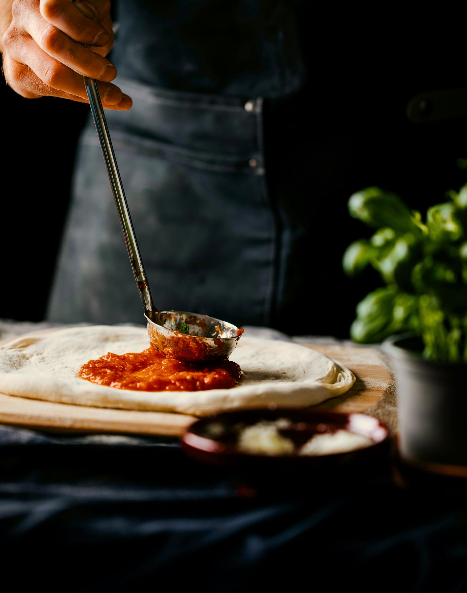 Ruka nanášajúca paradajkovú omáčku na cesto na pizzu s bylinkami v pozadí.