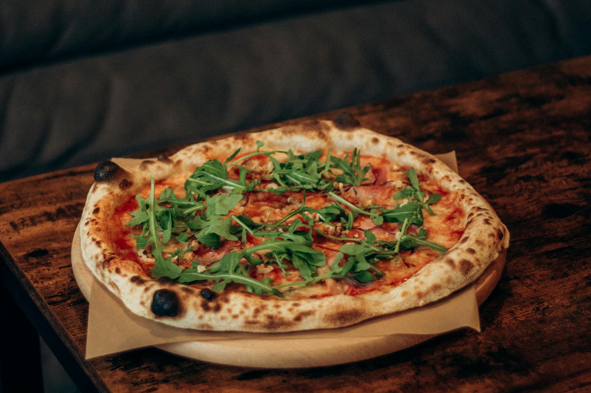 Pizza na drevenej doske so zelenou rukolou a tenkým chrumkavým okrajom, servírovaná na stole.