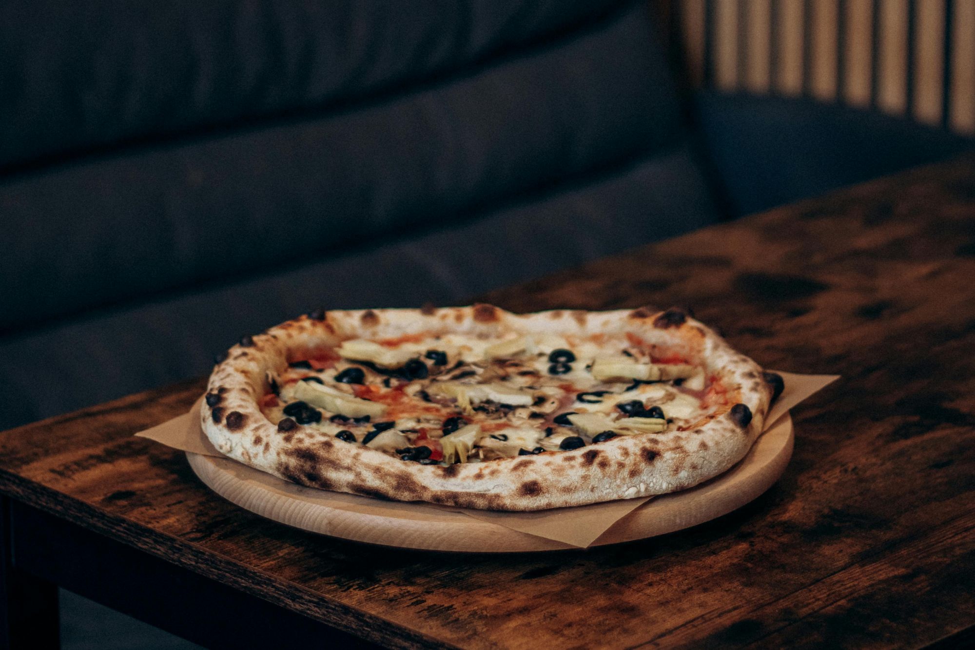 Pizza s olivami na drevenom tanieri, umiestnená na stole v útulnej kaviarni.