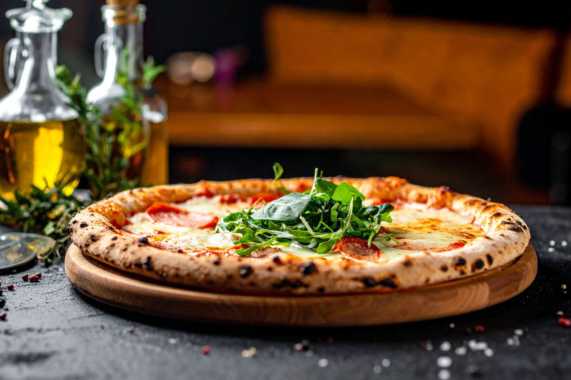 Pizza s mozarrelom, salaminom i svježim rukolom, poslužena na drvenoj dasci s uljem i začinima u pozadini.