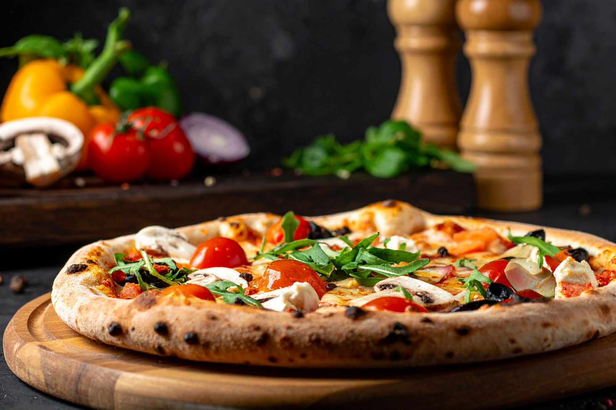 Pizaa s rajčicama, gljivama i svježim začinskim biljem na drvenoj dasci. U pozadini povrće i začini.
