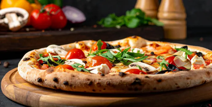 Pizaa s rajčicama, gljivama i svježim začinskim biljem na drvenoj dasci. U pozadini povrće i začini.