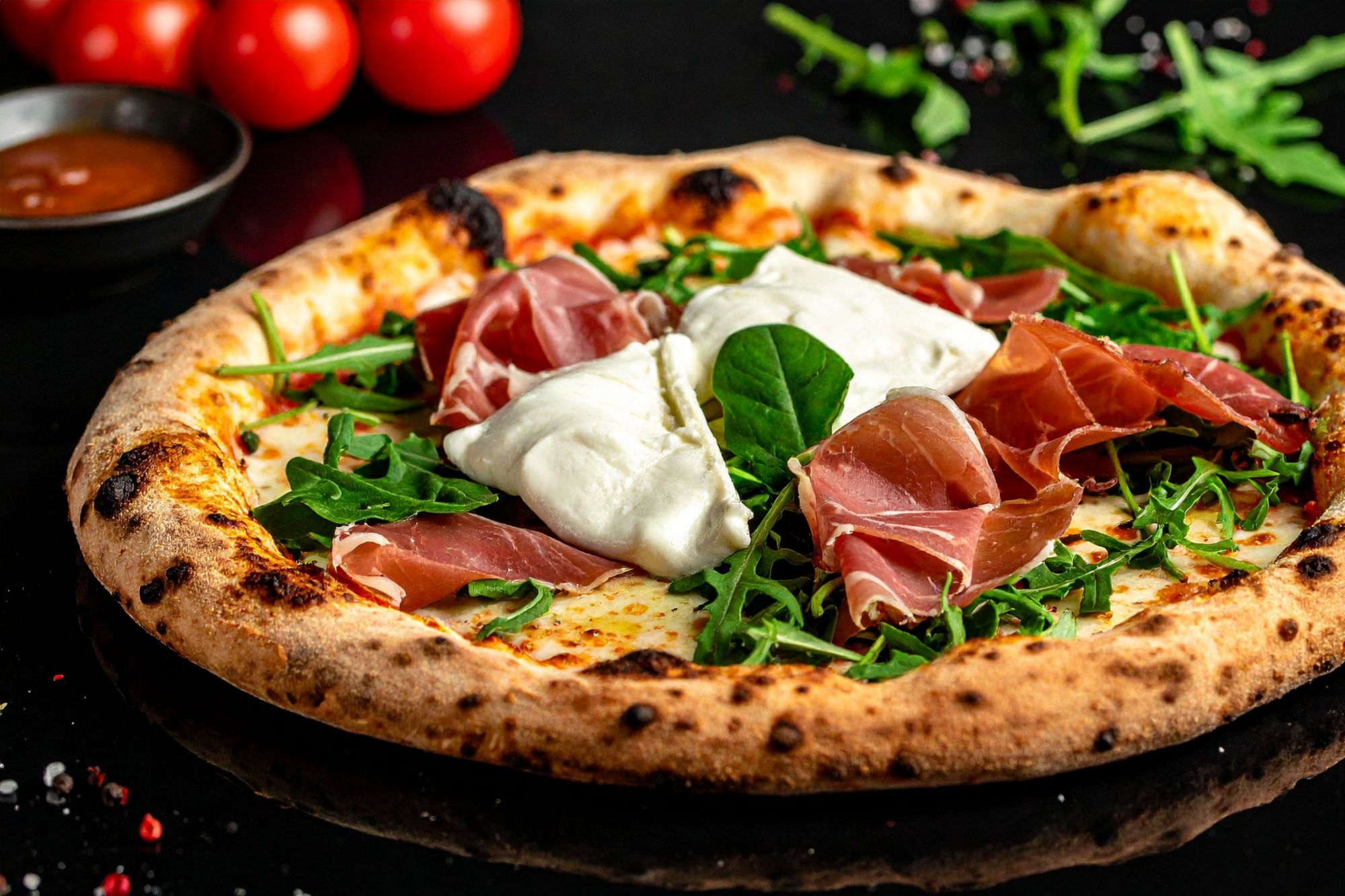 Pizza s pršutom, rukolom i mozzarellom, poslužena na crnoj podlozi uz svježe rajčice.