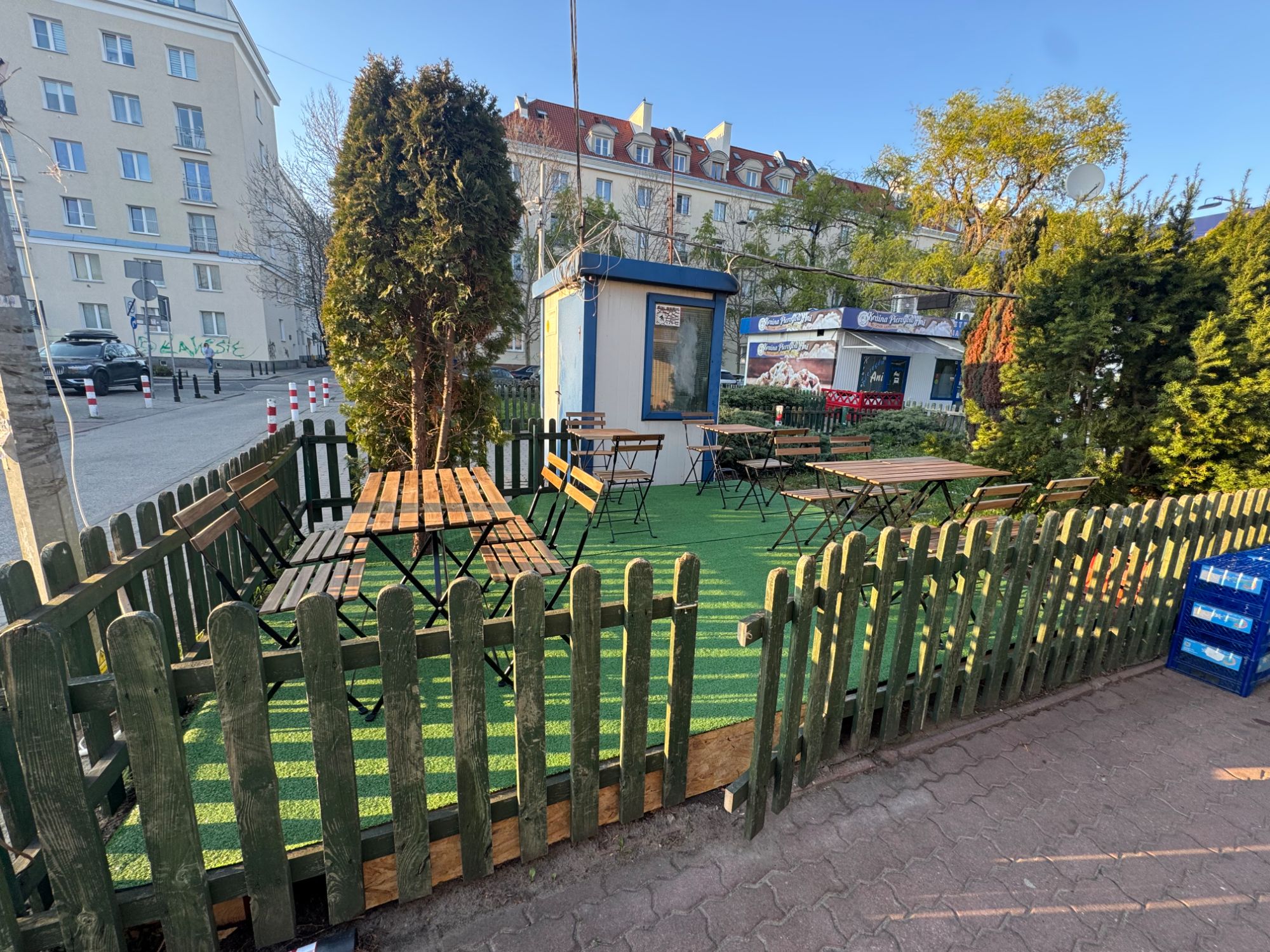Zielony ogródek z drewnianym płotem, stołami i krzesłami, otoczony drzewami, w miejskim otoczeniu.