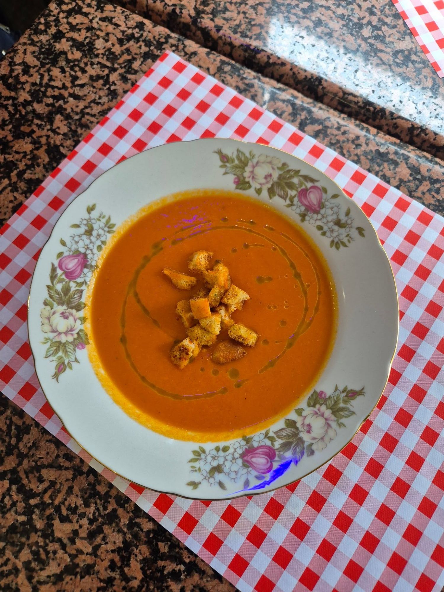 Zupa pomidorowa w białym talerzu z chrupiącymi dodatkami, podana na kratkowanym obruse.
