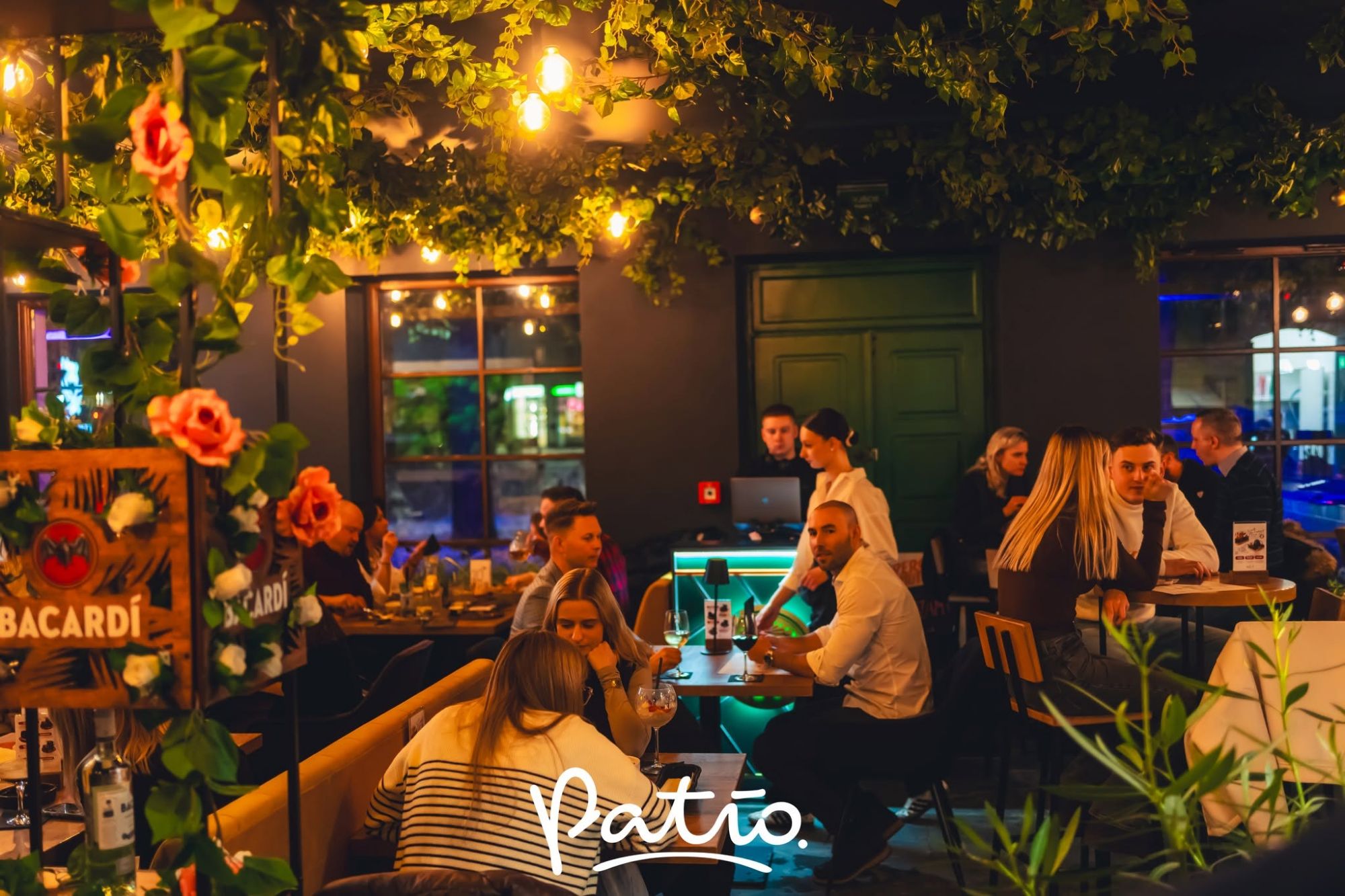 Ludzie w restauracji Patio przy lampach i dekoracjach roślinnych, cieszący się posiłkiem i towarzystwem.