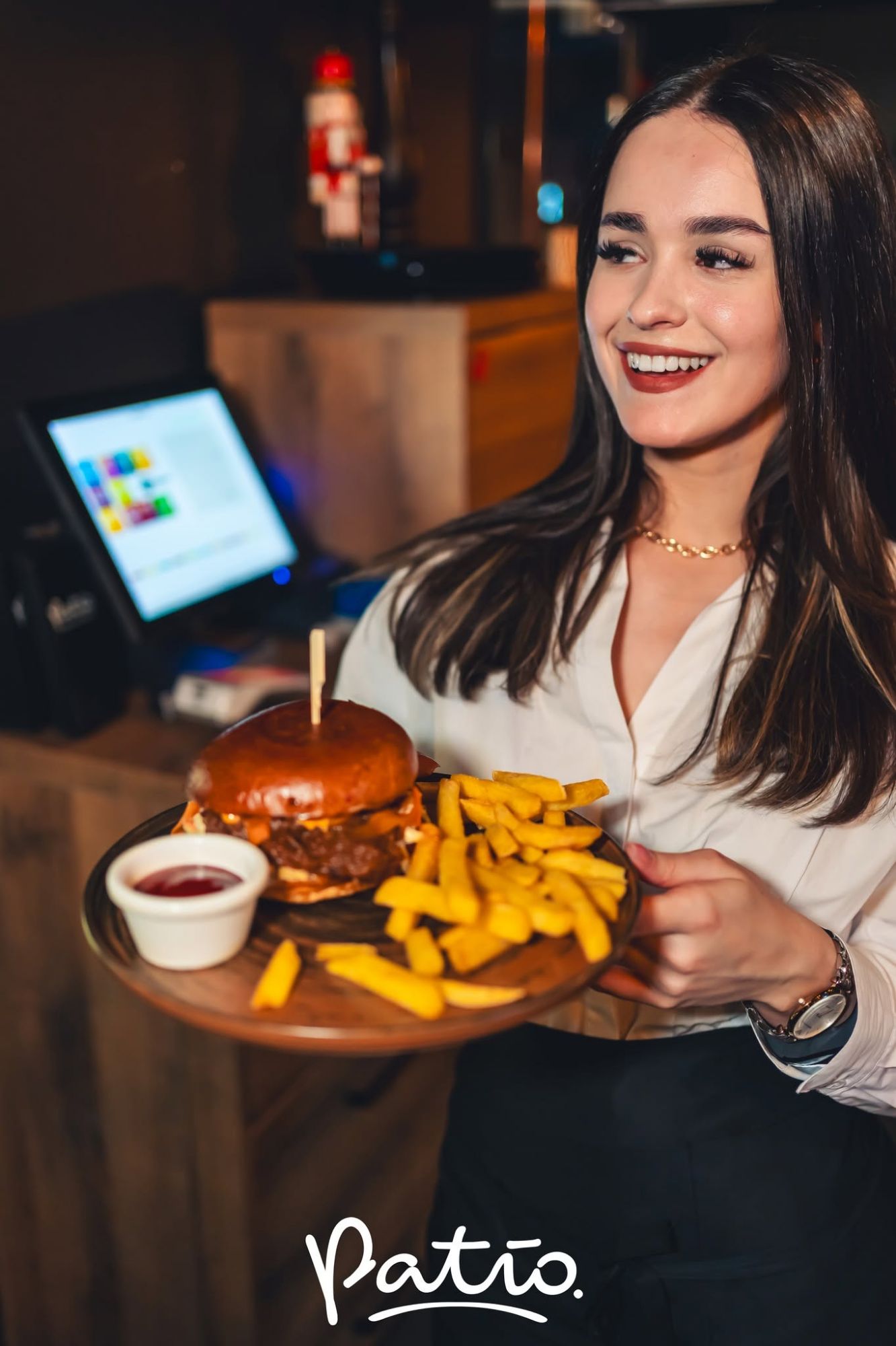 Młoda kobieta w białej koszuli trzyma talerz z burgerem i frytkami w restauracji.