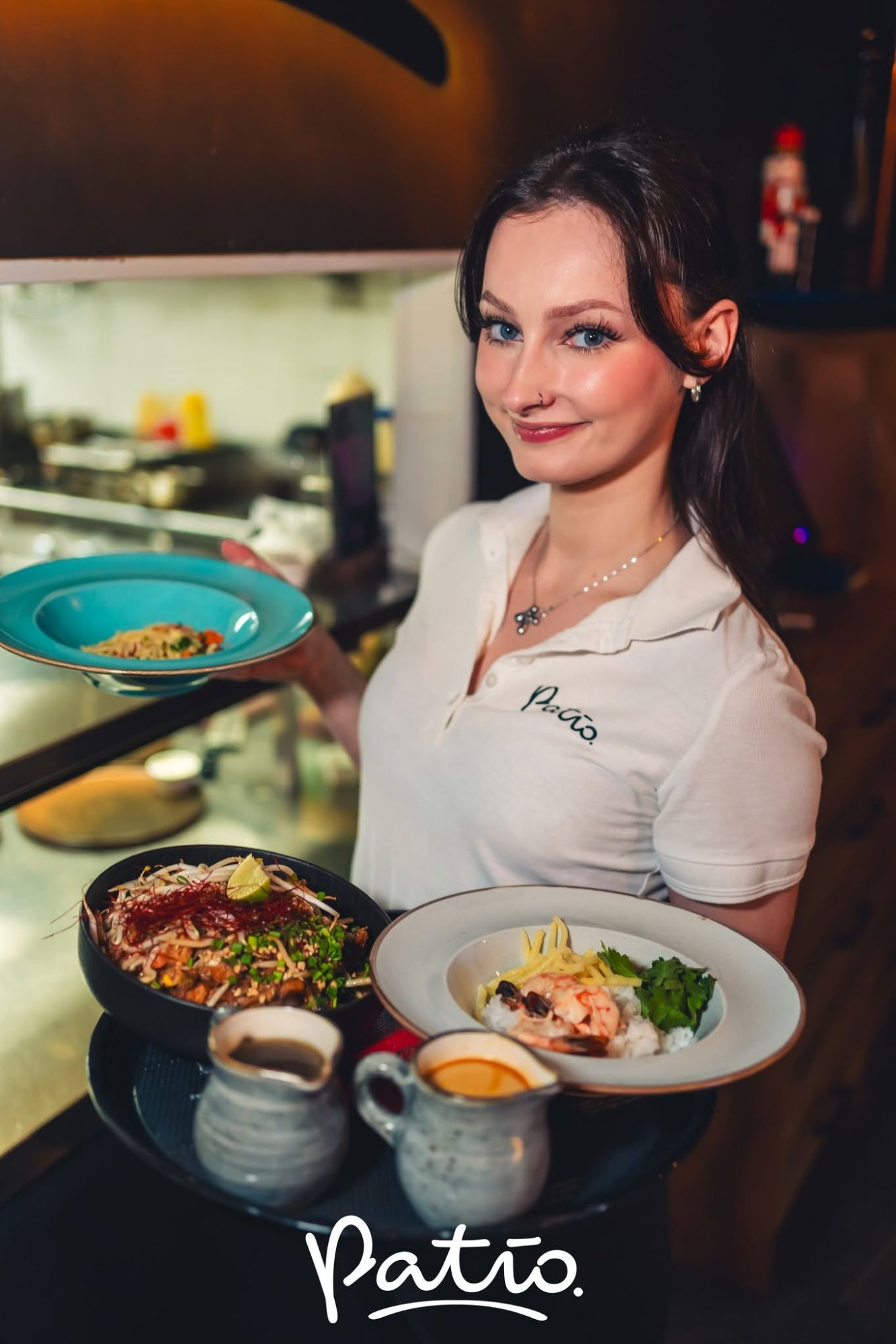 Kobieta w białej koszulce serwuje dania w restauracji Patio, z kuchnią w tle.