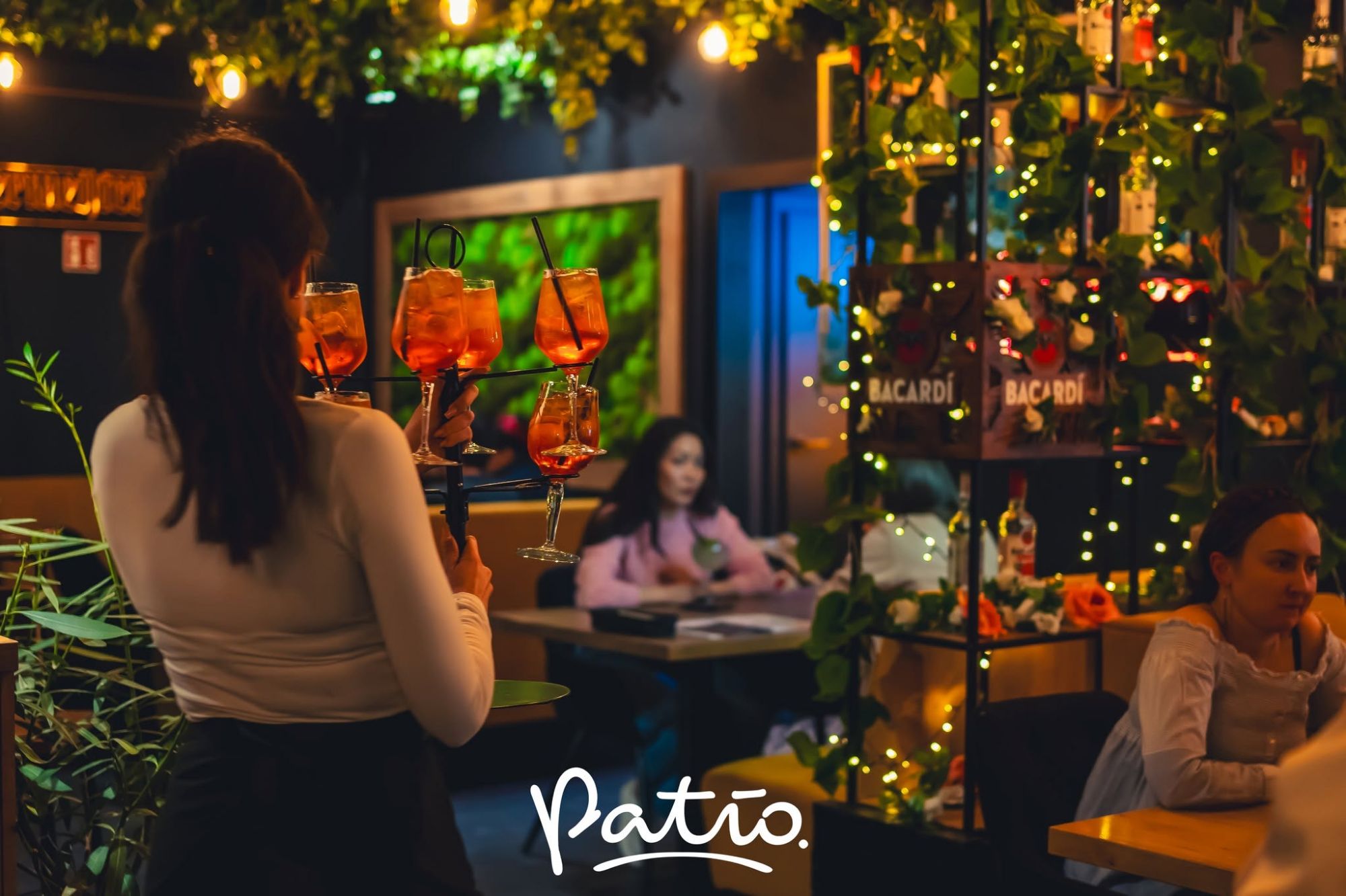 Kelnerka podaje drinki w stylowej restauracji Patio, z oświetleniem i roślinami w tle.