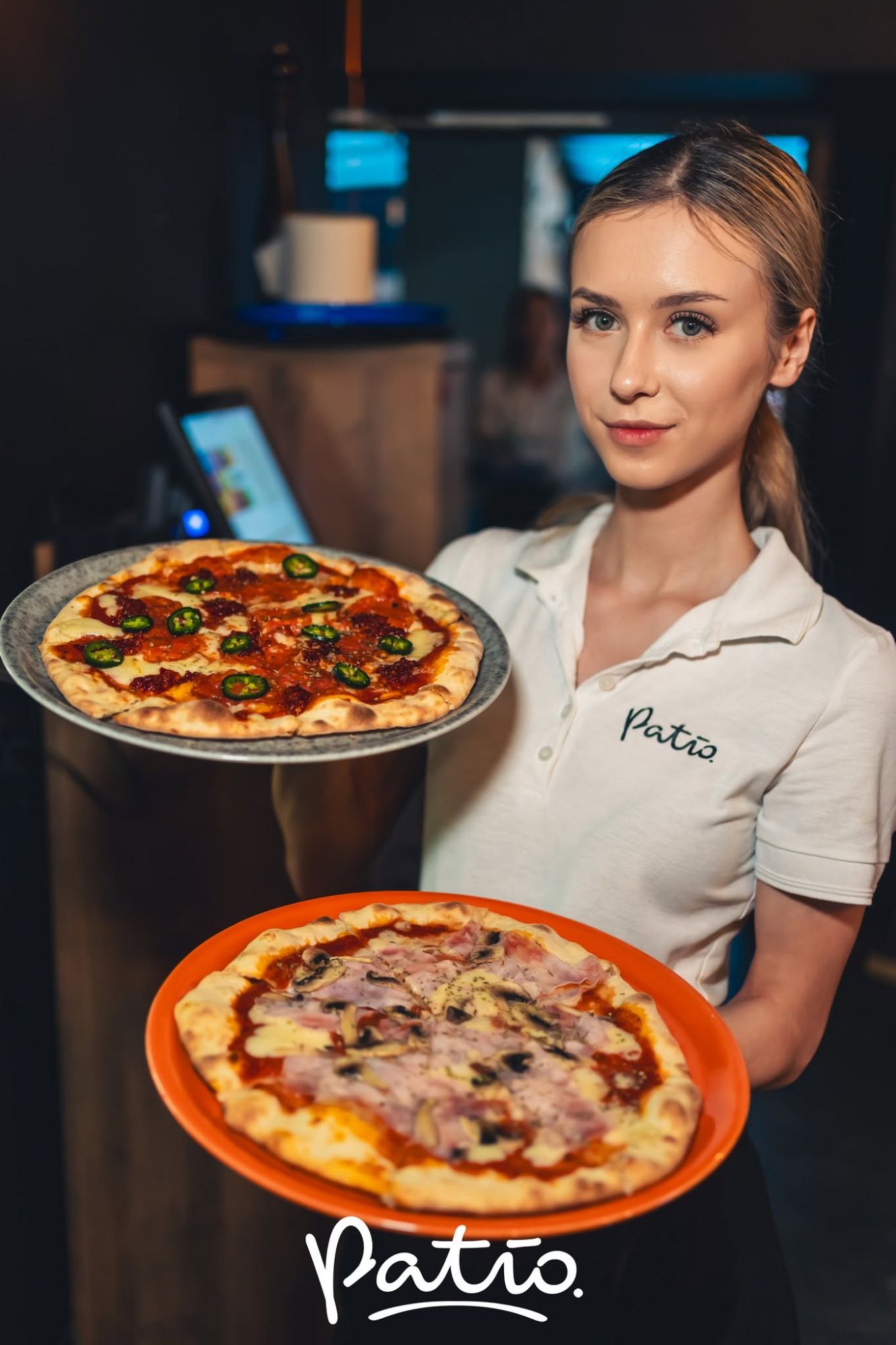 Kelnerka trzyma dwie pizze w restauracji Patio, jedna z papryczkami jalapeño, druga z szynką i oliwkami.