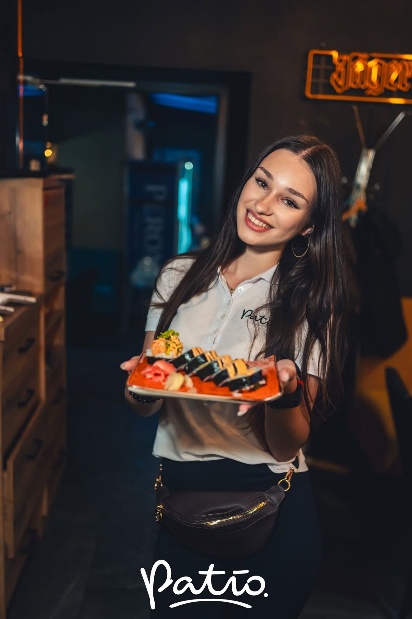 Młoda kobieta w koszulce z logo "Patio" trzyma talerz sushi w restauracji.
