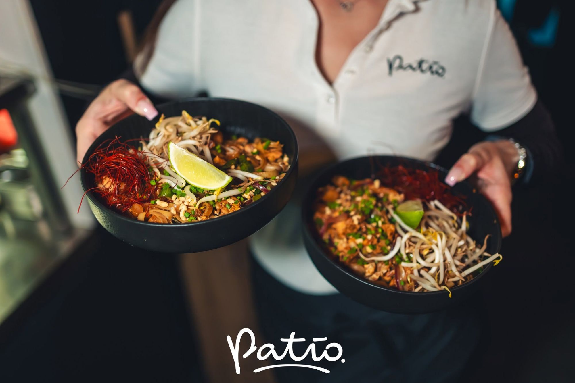 Osoba trzymająca dwa czarne miski z jedzeniem, udekorowane ziołami i limonką, w restauracji Patio.