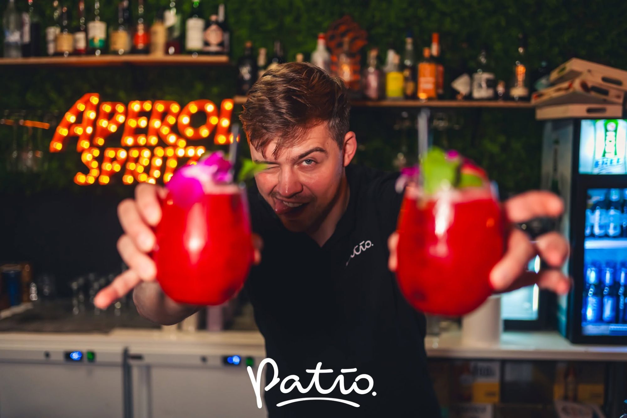 Barman trzyma dwa kolorowe koktajle, z napisem "Patio" w dolnej części i oświetleniem w tle.