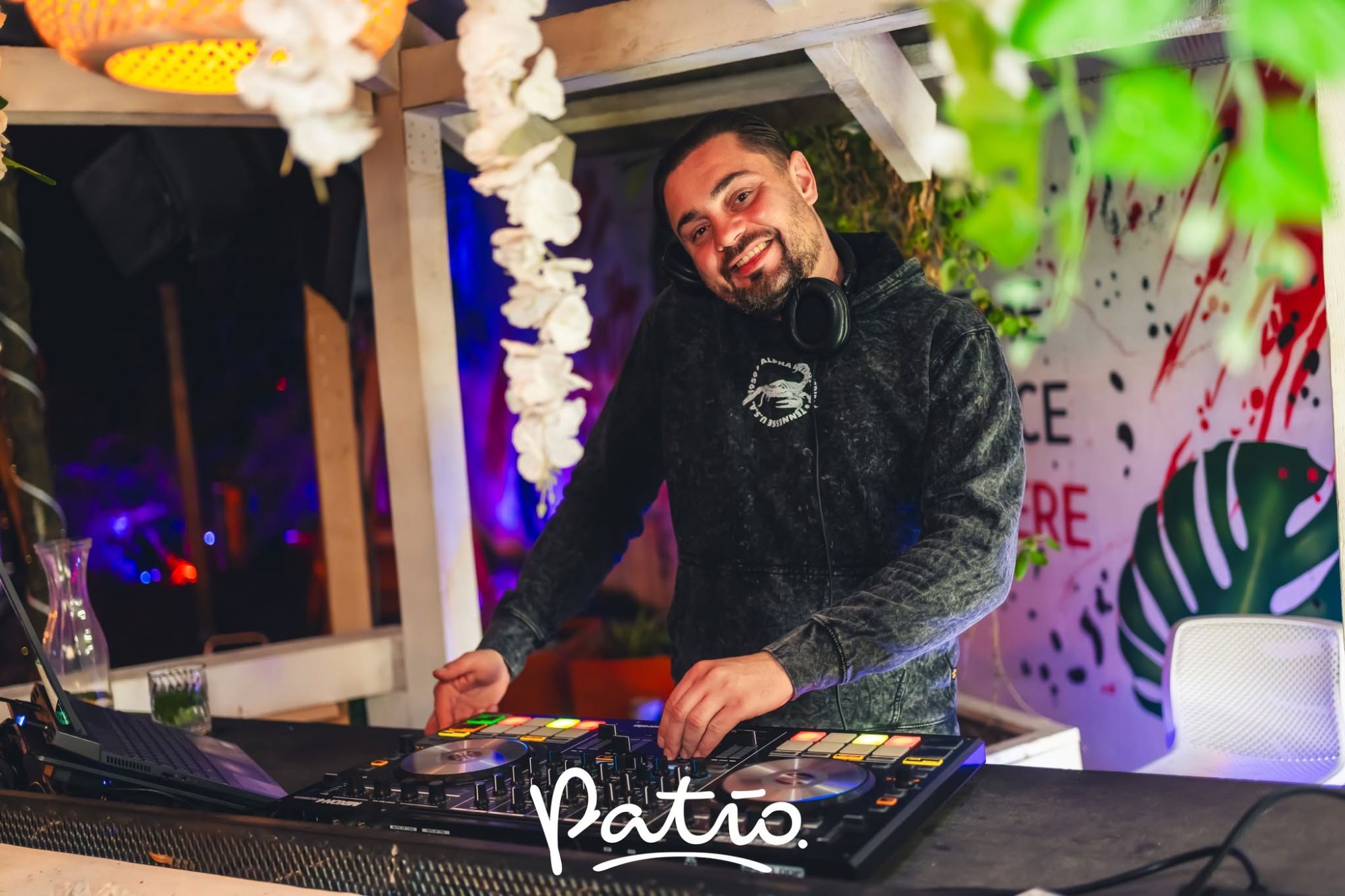 DJ w klubie, grający na konsoli, z uśmiechem i słuchawkami, otoczony przez roślinność i kolorowe światła.