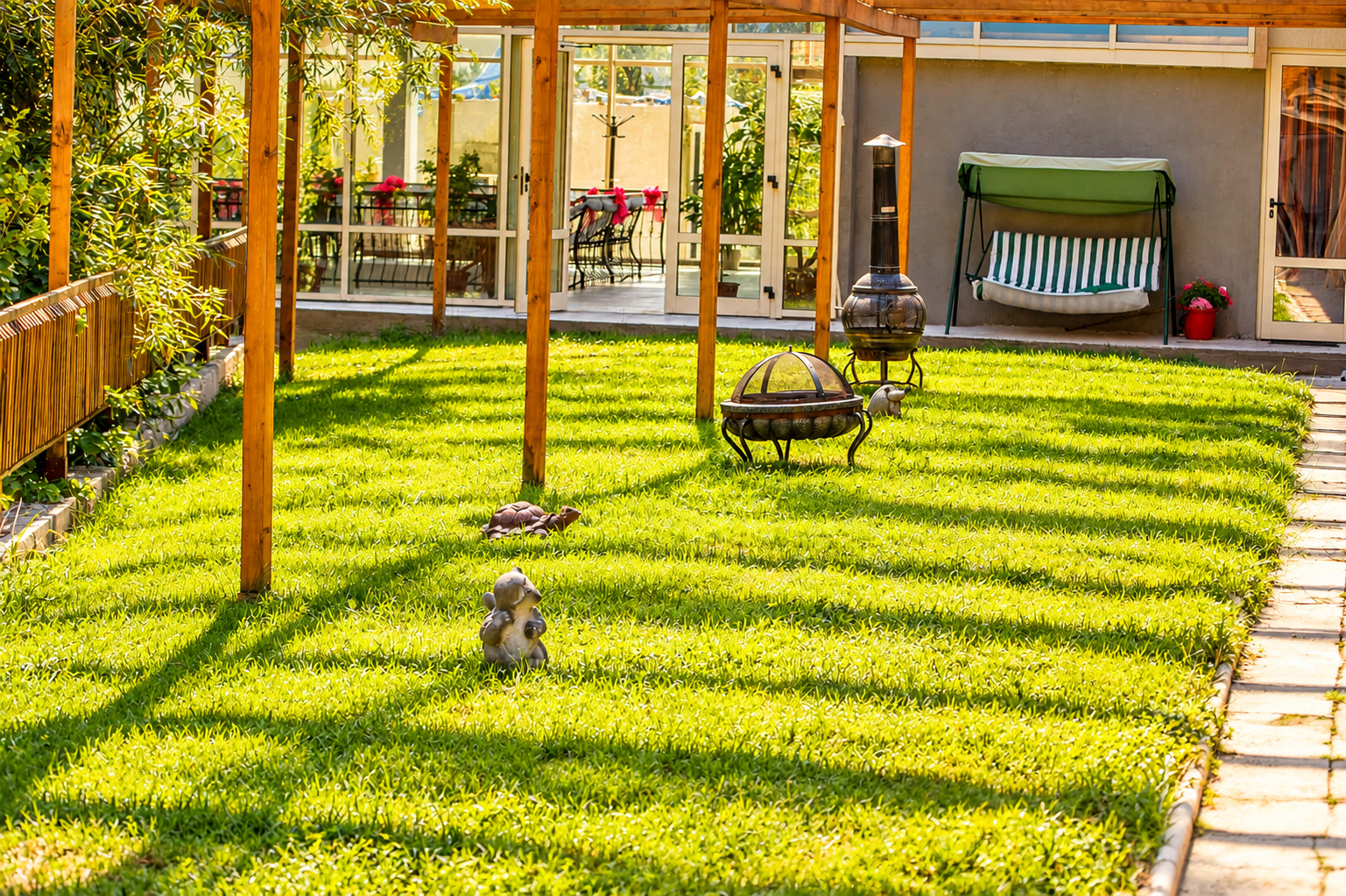 Grădină cu iarbă verde, structură de lemn, statui decorative și mobilier de grădină la soare.