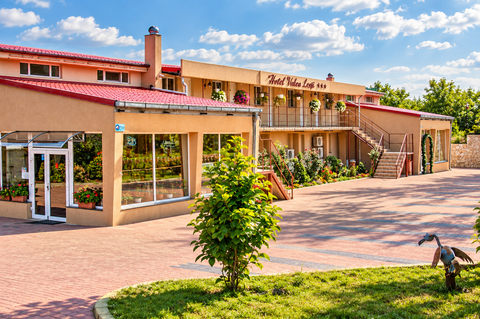 Hotel frumos cu fațadă roz, flori, curte spațioasă și cer senin, situat într-o zonă liniștită.