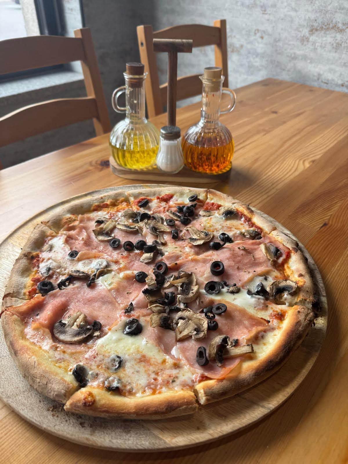 Pizza z szynką, czarnymi oliwkami i pieczarkami na drewnianej desce, w tle oliwy w dekoracyjnych butelkach.