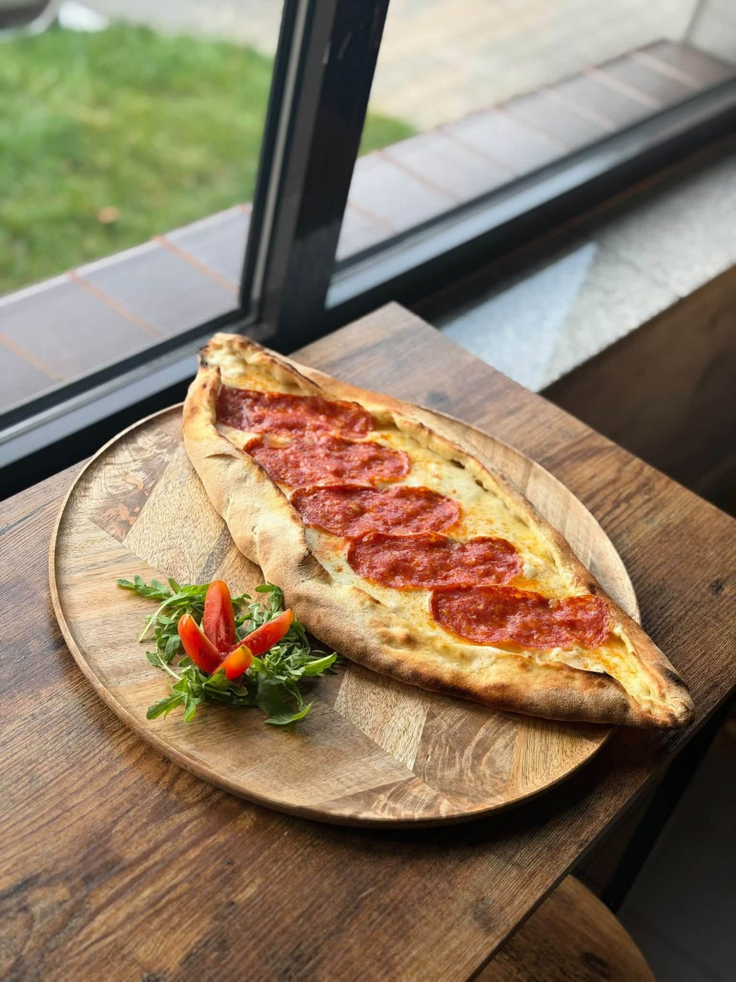 Pizza z pepperoni na talerzu z sałatą i pomidorem, umieszczona przy oknie.