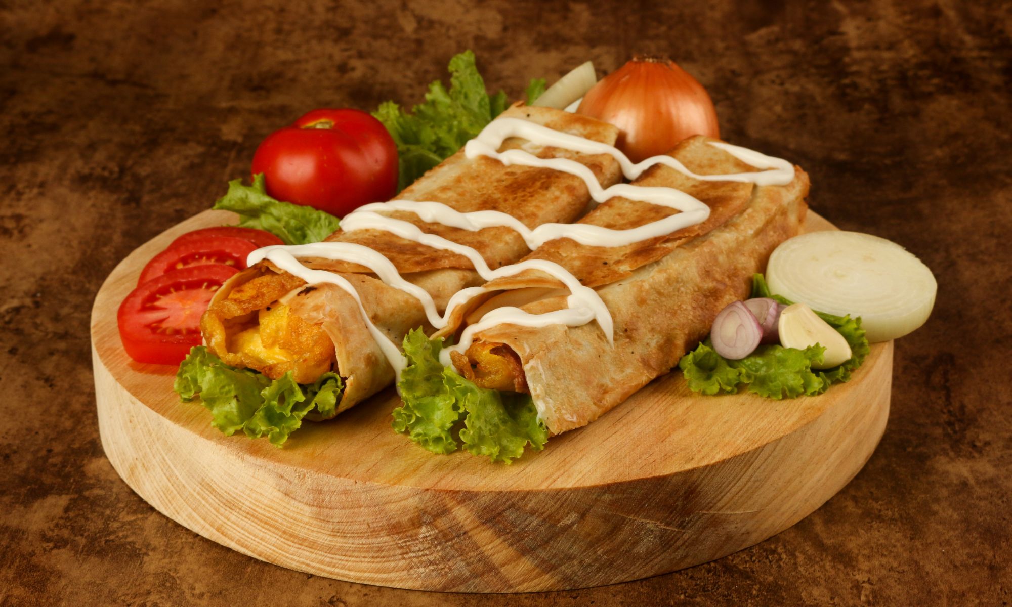 Dwa smaczne burrito na drewnianej desce, podane z sałatą, pomidorami i cebulą, polane sosem.